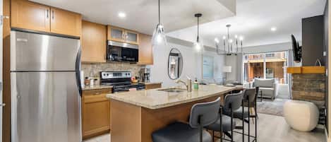 A modern kitchen with a stainless steel refrigerator, wooden cabinets, granite countertops, and a central island with bar stools.