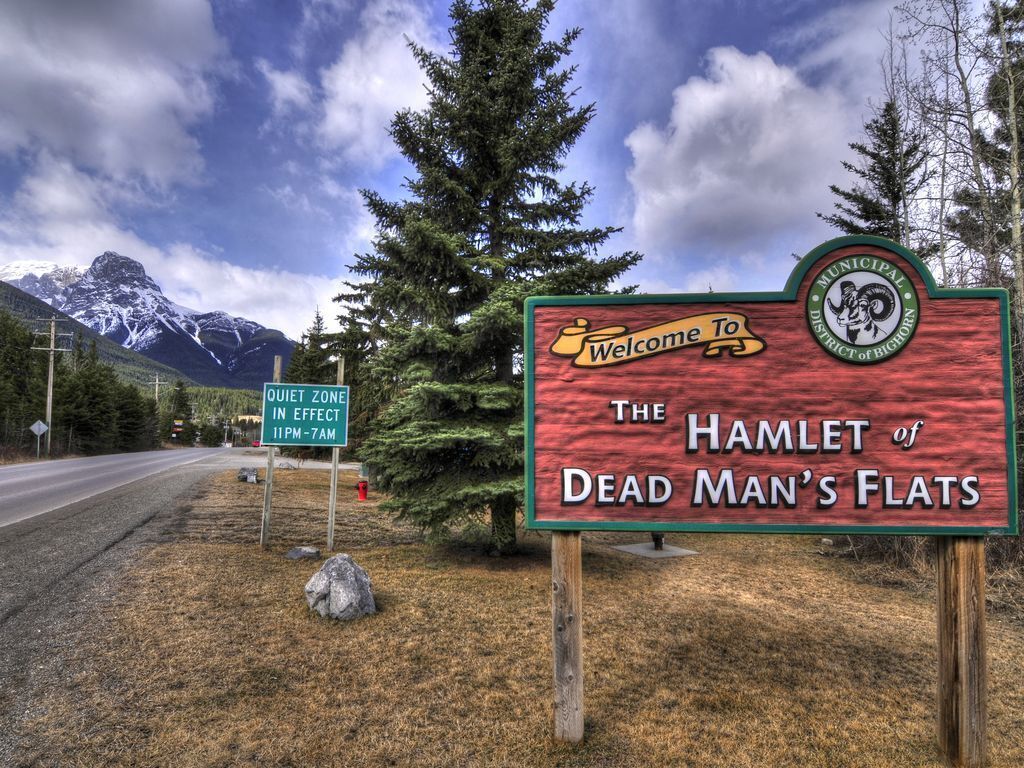 A sign welcoming visitors to the Hamlet of Dead Man's Flats with a mountainous backdrop.