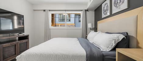 A modern bedroom with a bed, a TV on a wooden cabinet, a window with curtains, and framed artwork on the wall.