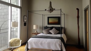 A bedroom with a four-poster bed, a bedside lamp, a framed picture on the wall, and a large window with a view of a brick building.