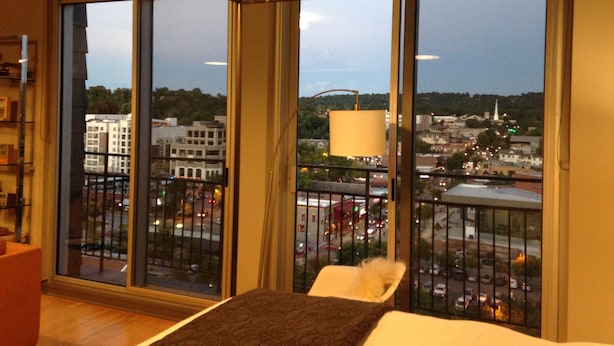 A hotel room with large windows, a bed, a chair, and a lamp.