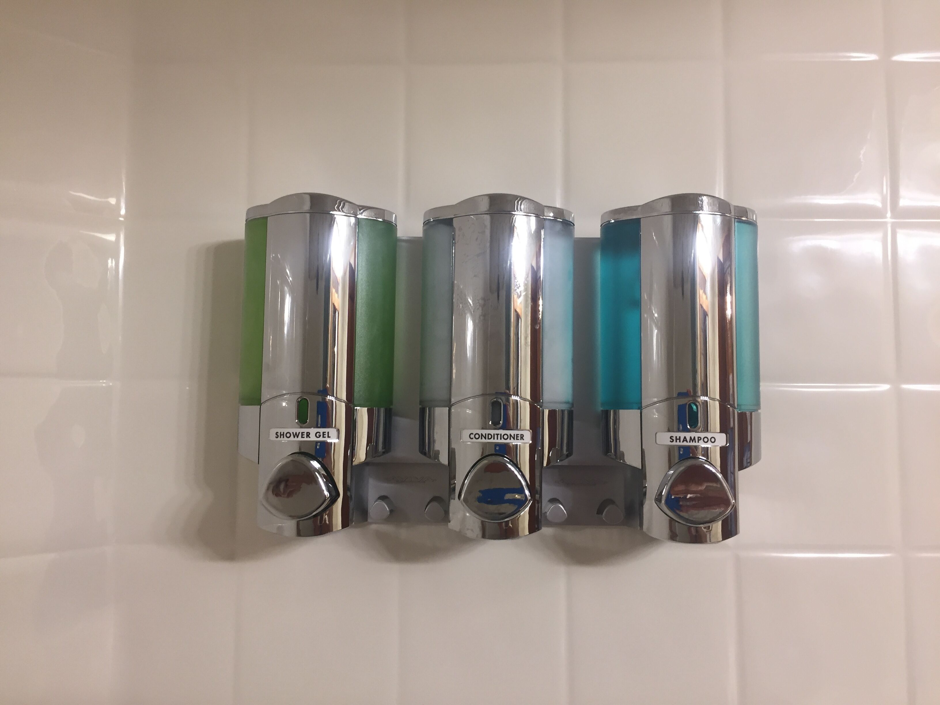 Dispensador mural con tres compartimentos etiquetados para gel de ducha, acondicionador y champú.