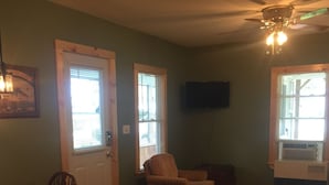 A living room with a dining area, wooden flooring, a ceiling fan, and a television mounted on the wall. - Year Round Lakeview Cottage with Dock on Lake Champlain (Alburg)