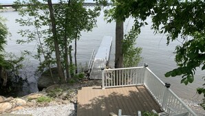 A wooden deck with a white railing overlooking a calm body of water, surrounded by trees and rocks. - Year Round Lakeview Cottage with Dock on Lake Champlain (Alburg)
