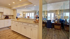 A spacious kitchen with white cabinets, a granite countertop, and a large island. There is a dining area with a blue sofa and a mirror reflecting the kitchen. - Breathtaking Penthouse w/ Direct Oceanfront Views,  40' private balcony. (Daytona Beach Shores)