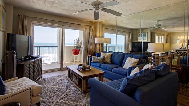 A living room with a large blue sofa, a coffee table, a TV, and a balcony with a view of the ocean. - Breathtaking Penthouse w/ Direct Oceanfront Views,  40' private balcony. (Daytona Beach Shores)