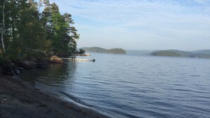 A beach with a dock extending into calm waters, surrounded by trees and distant hills.