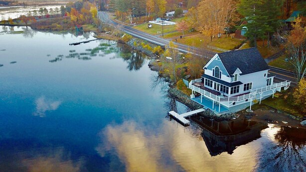 A house with a dock extending into a calm lake, surrounded by trees and a road.