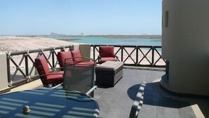 Een balkon met rode stoelen en een glazen tafel, met uitzicht op een strand.