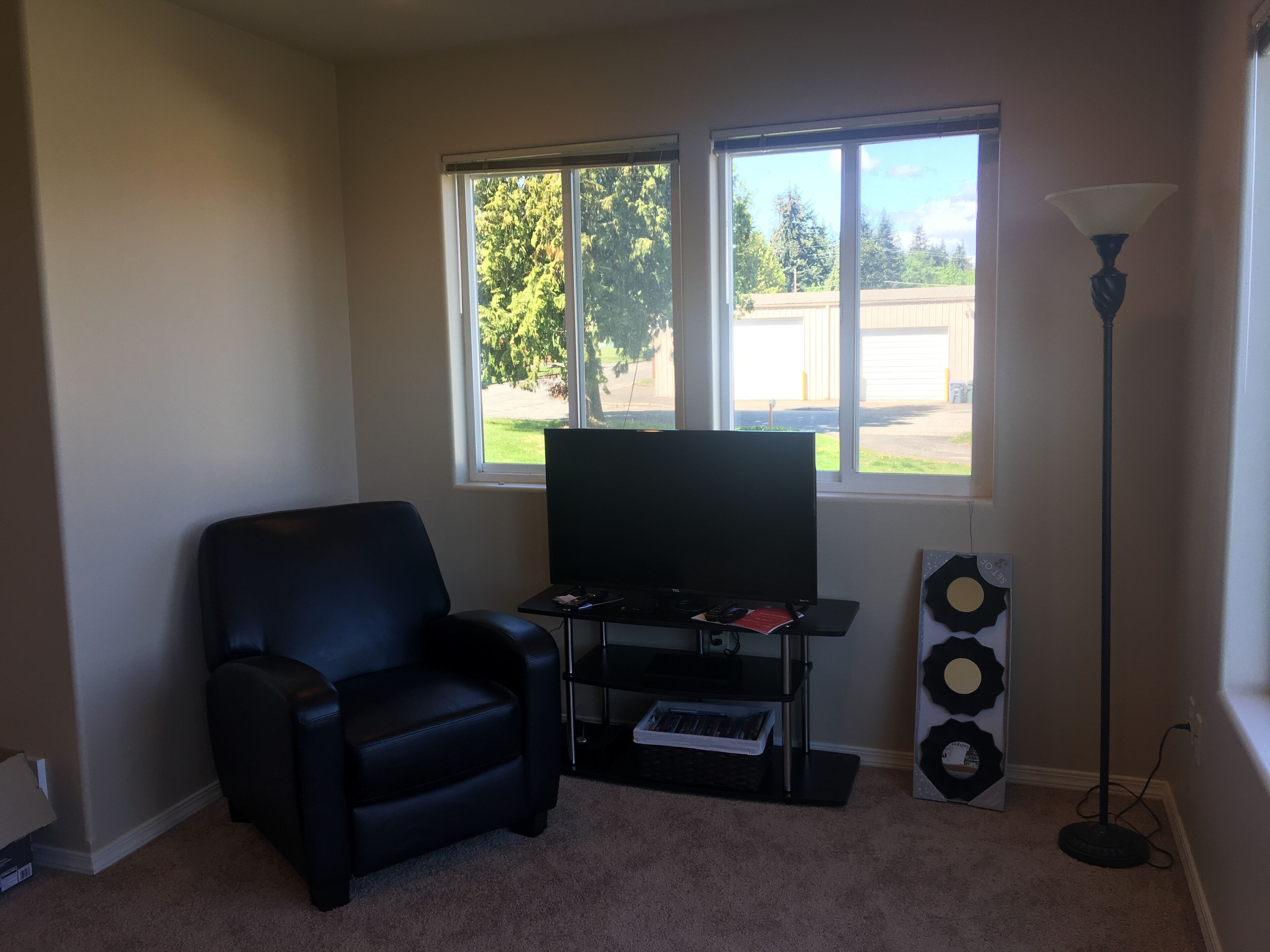 A living room with a black leather chair, a flat-screen TV on a stand, a floor lamp, and a window with a view of a backyard.