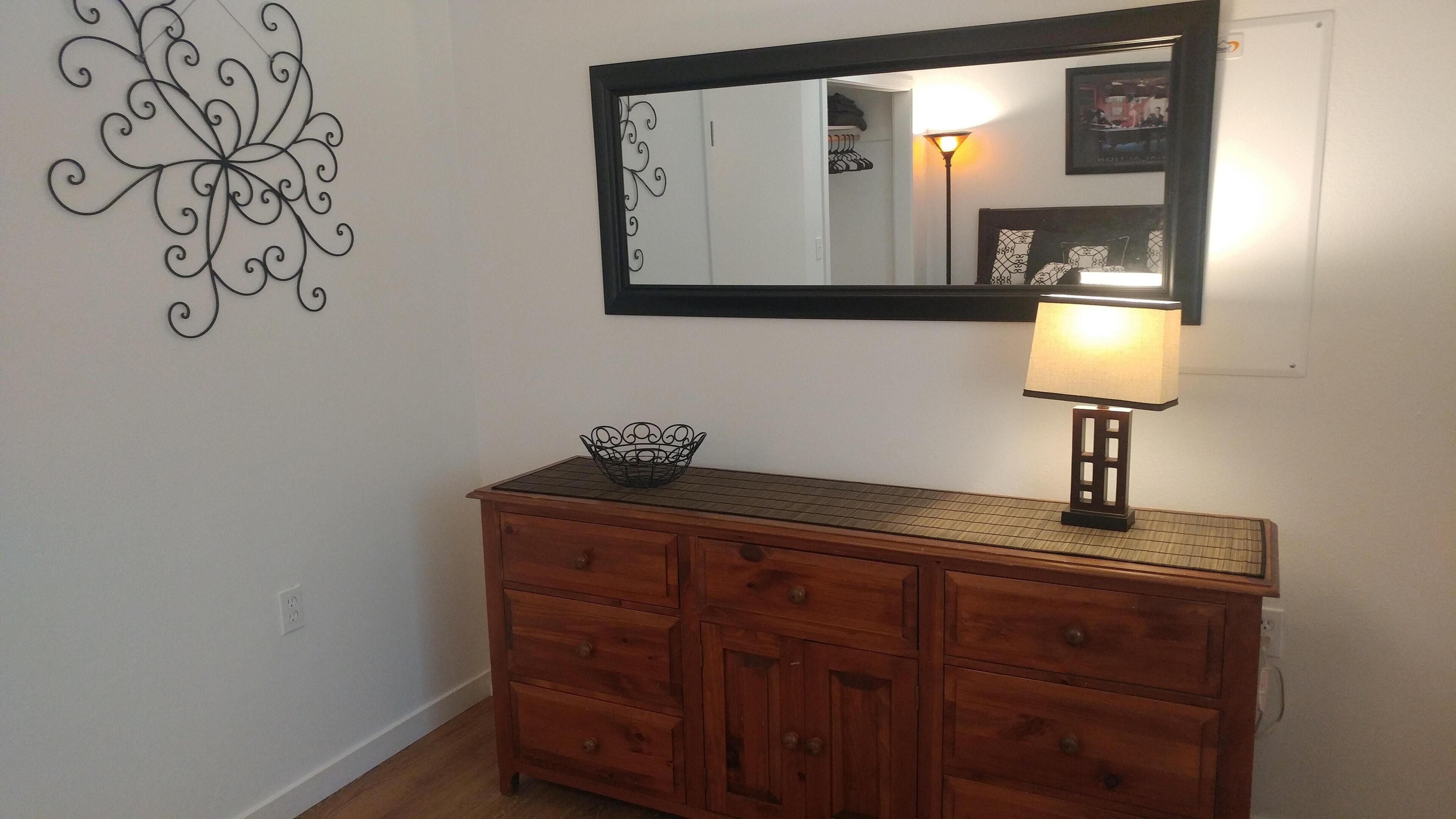 A wooden dresser with a mirror above it, a lamp on the dresser, and a decorative metal wall piece.
