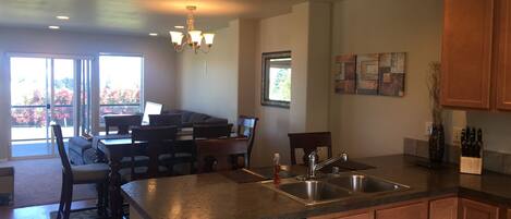A modern kitchen with a dishwasher, double sink, and wooden cabinets. There is a dining area with a table and chairs, and a large window providing natural light.