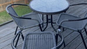 A round table with four chairs on a wooden deck, overlooking a fenced yard with trees and houses in the background. - Mountains to Sea Twin (Port Angeles)