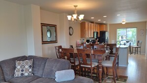 A spacious living room with a gray sofa, wooden dining table, and chairs. A kitchen with wooden cabinets and a refrigerator is visible in the background. - Mountains to Sea Twin (Port Angeles)