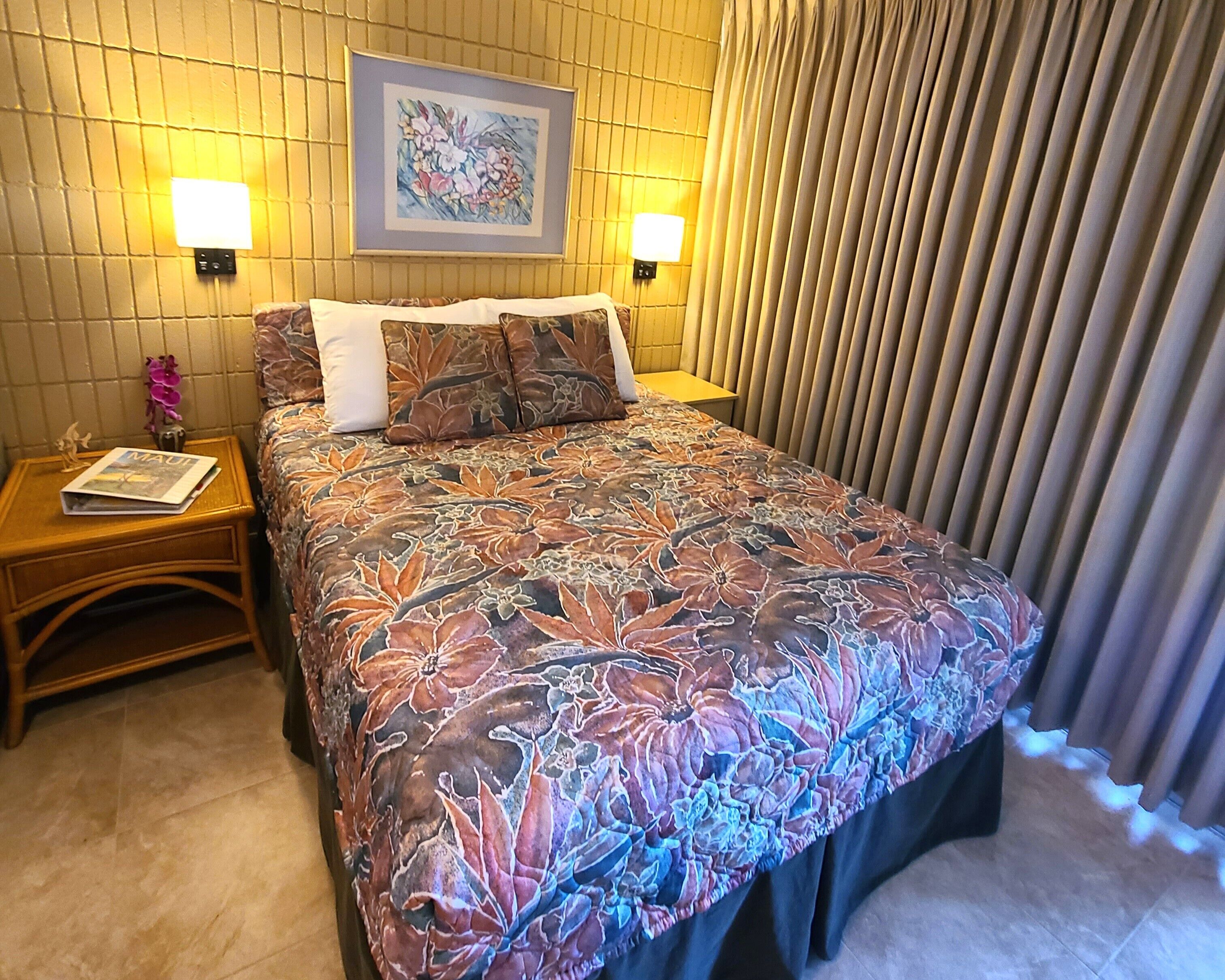 Ett hotellrum med ett blommigt överkast på sängen, två sänglampor, ett inramat konstverk ovanför sängen och en gardin vid fönstret.