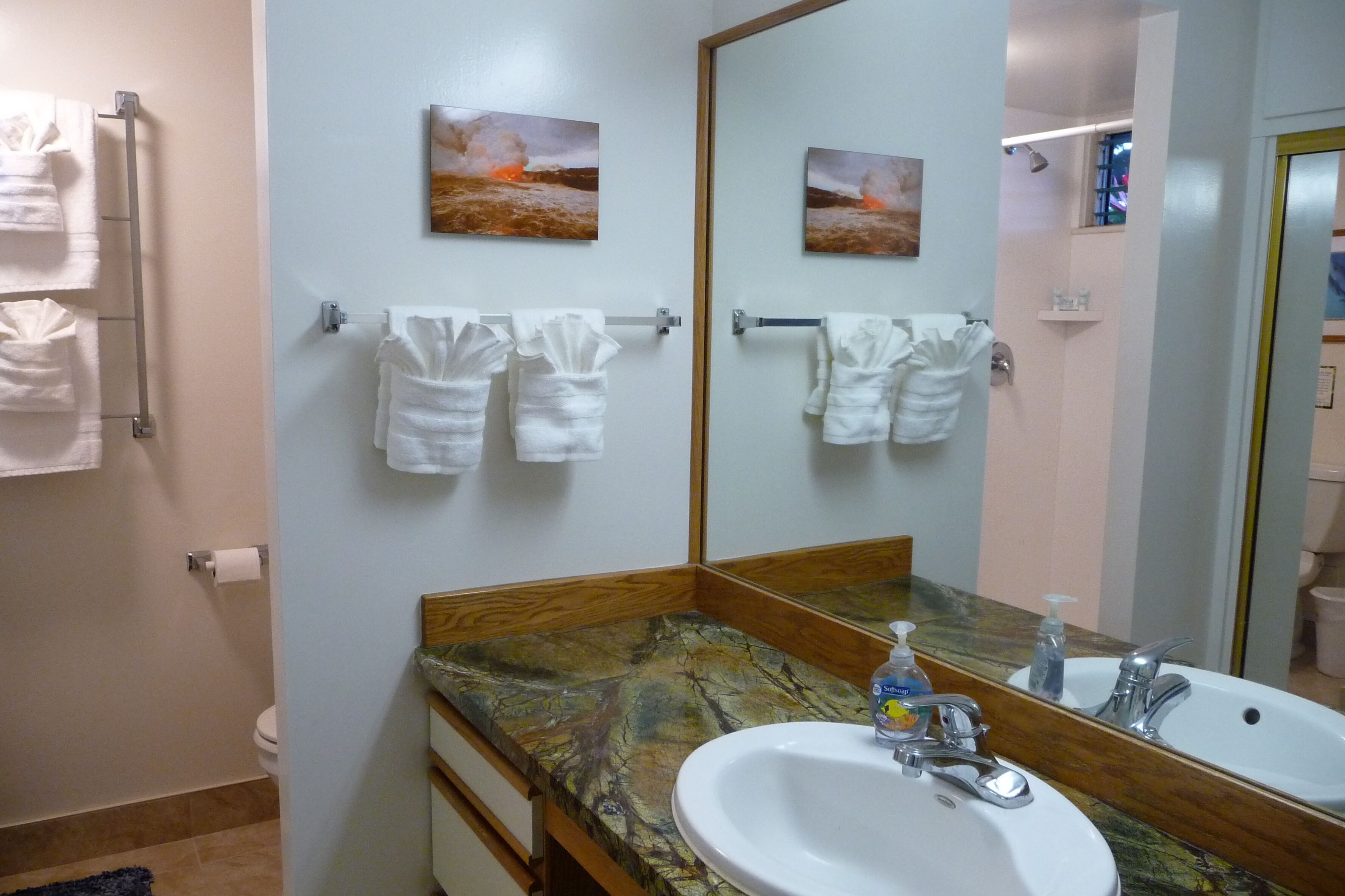Une salle de bain avec un comptoir en marbre, un lavabo blanc et un grand miroir. Des serviettes sont suspendues sur un porte-serviettes et un tableau représentant une scène de plage est accroché au mur.