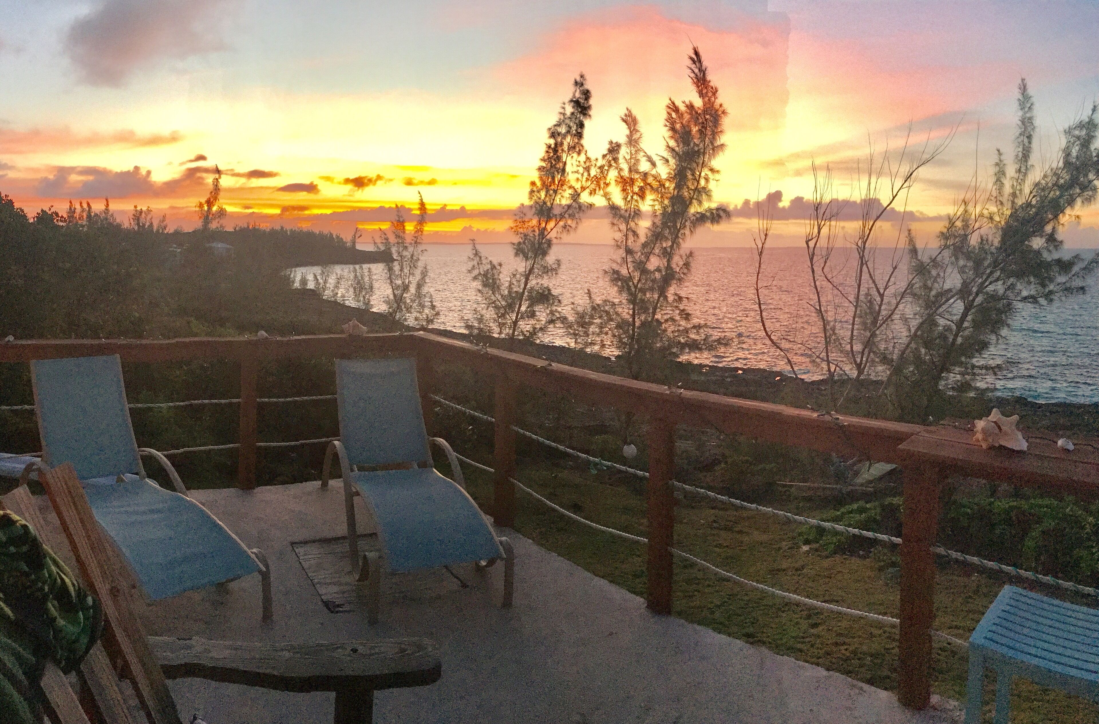 Eine Terrasse mit zwei Liegestühlen und einem kleinen Tisch, mit Blick auf ein Gewässer und einen Sonnenuntergang.