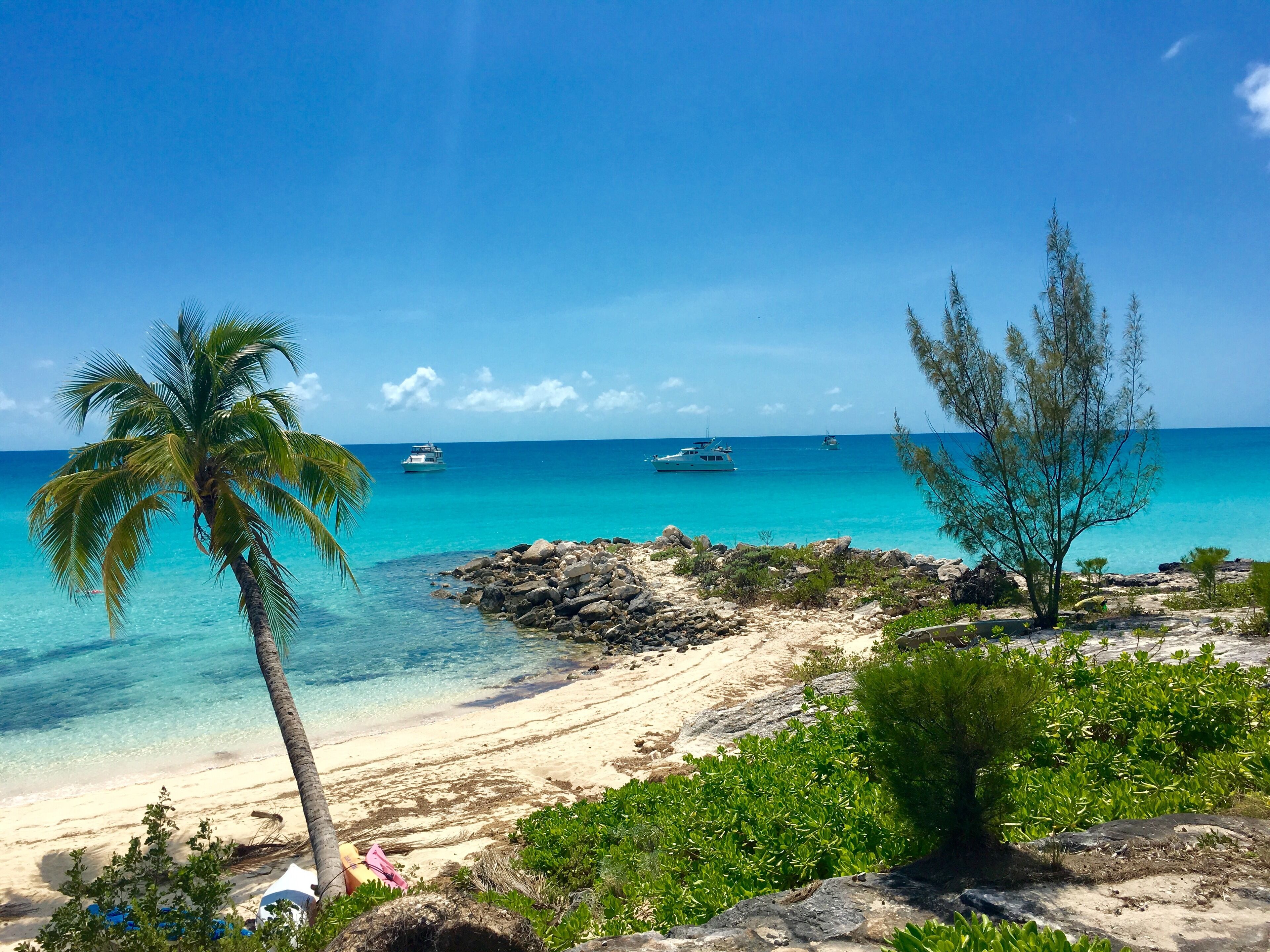 Une plage à l’eau turquoise et limpide, un rivage sablonneux et quelques bateaux visibles au loin.