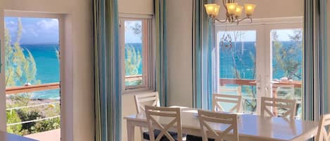 A dining area with a table and chairs, large windows with curtains, and a view of the ocean.