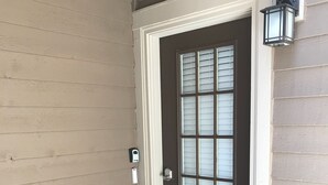 A front door with a dark panel and a window with blinds, a small porch with a doormat, and a wall-mounted lantern.