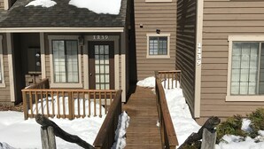 A residential building with a wooden deck leading to the entrance, surrounded by snow.