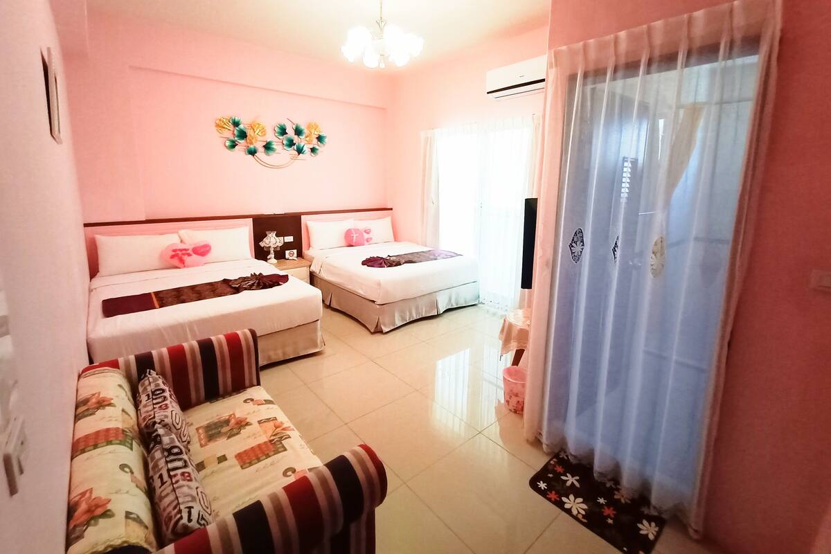 A hotel room with two beds, a sofa, a chandelier, and a window with curtains.