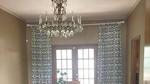 Ein Esszimmer mit Holztisch und -stühlen, einer Kristallleuchtre, gemusterten Vorhängen und einer Topfpflanze.