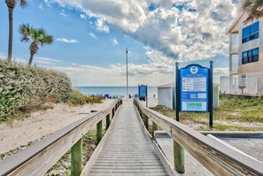 A wooden boardwalk leading to beach access, with a safety sign and a building on the right.