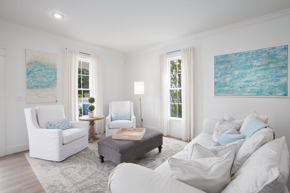 A spacious living room with a white sofa, a grey ottoman, and a white armchair. There are two windows with curtains, a round table with a lamp, and a large abstract painting on the wall.