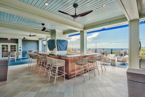 A bar area with a wooden bar and wicker chairs, a ceiling fan, and a view of the ocean.