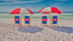 Zwei Liegestühle mit roten und weißen Sonnenschirmen an einem Sandstrand, im Hintergrund Brandungs Wellen.