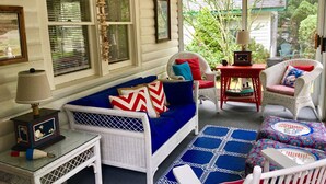 Une pièce chaleureuse avec un canapé bleu, des fauteuils en osier, une petite table avec une lampe et une vue sur la verdure à l’extérieur.