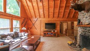 Interior acogedor de una cabaña con techo de madera, chimenea de piedra, cabeza de venado montada, televisor y sofá de cuero.