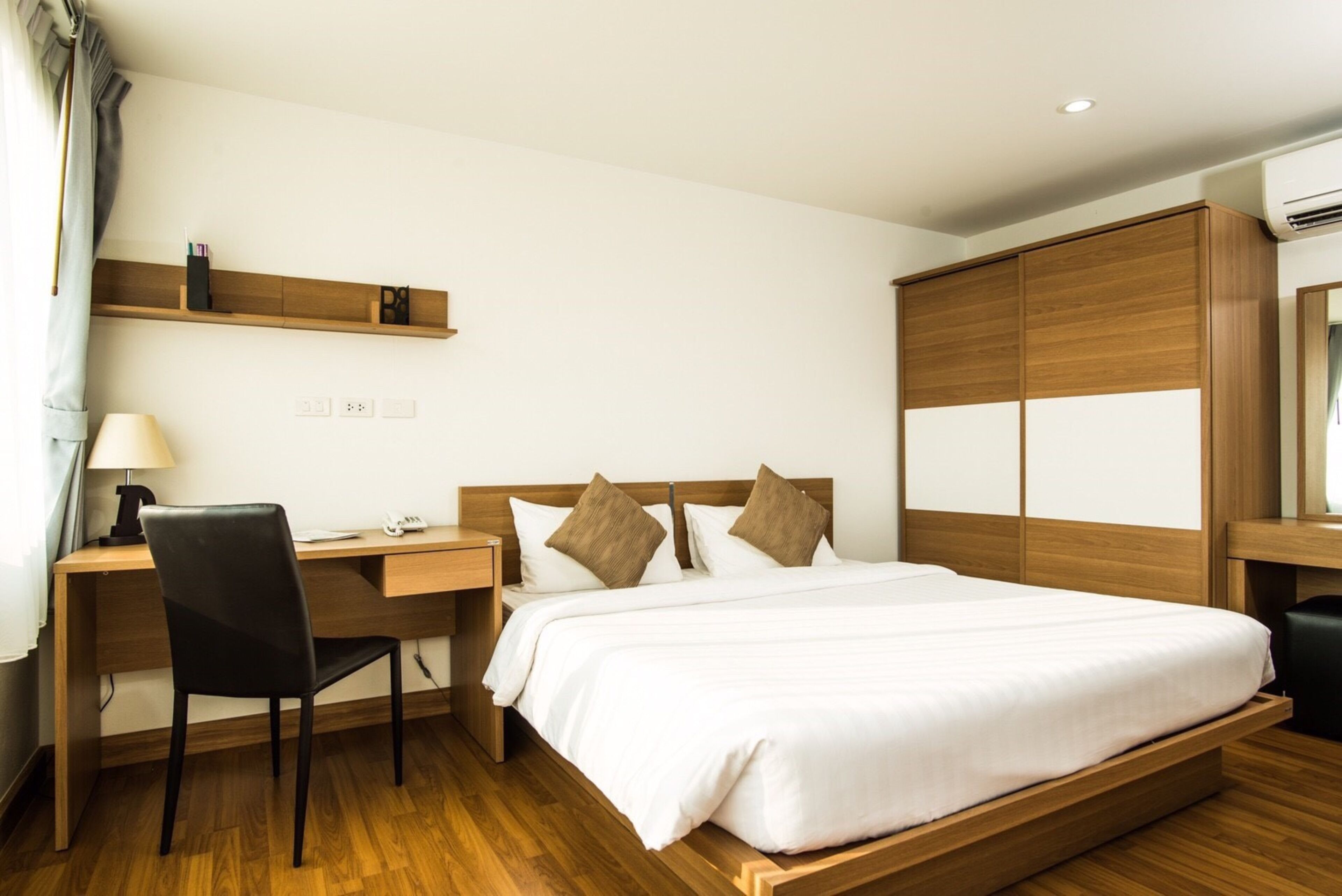 A hotel room with a wooden bed, a desk with a chair, a lamp, and a shelf with items.