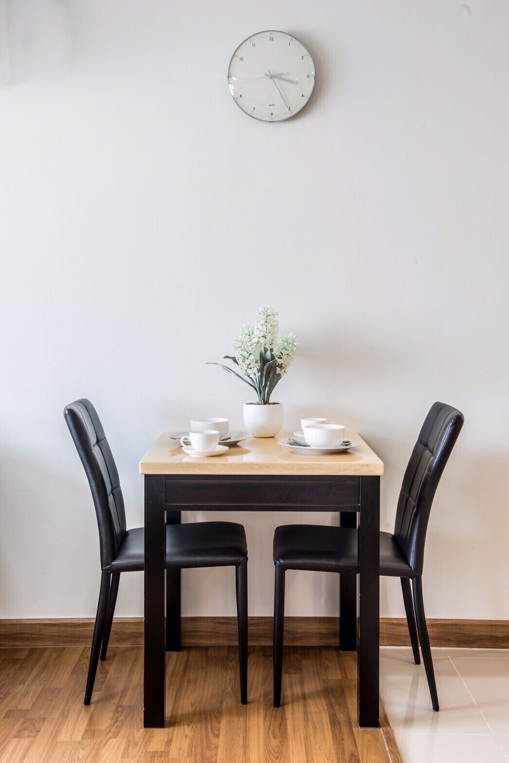 Una mesa de comedor con dos sillas negras, una planta en maceta y tazas y platillos blancos sobre la mesa.