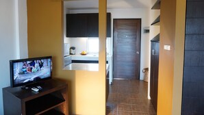 A living room with a television on a wooden stand, a kitchen area with cabinets and a window, and a hallway leading to a door. - Phompassorn Apartment (Chalong)