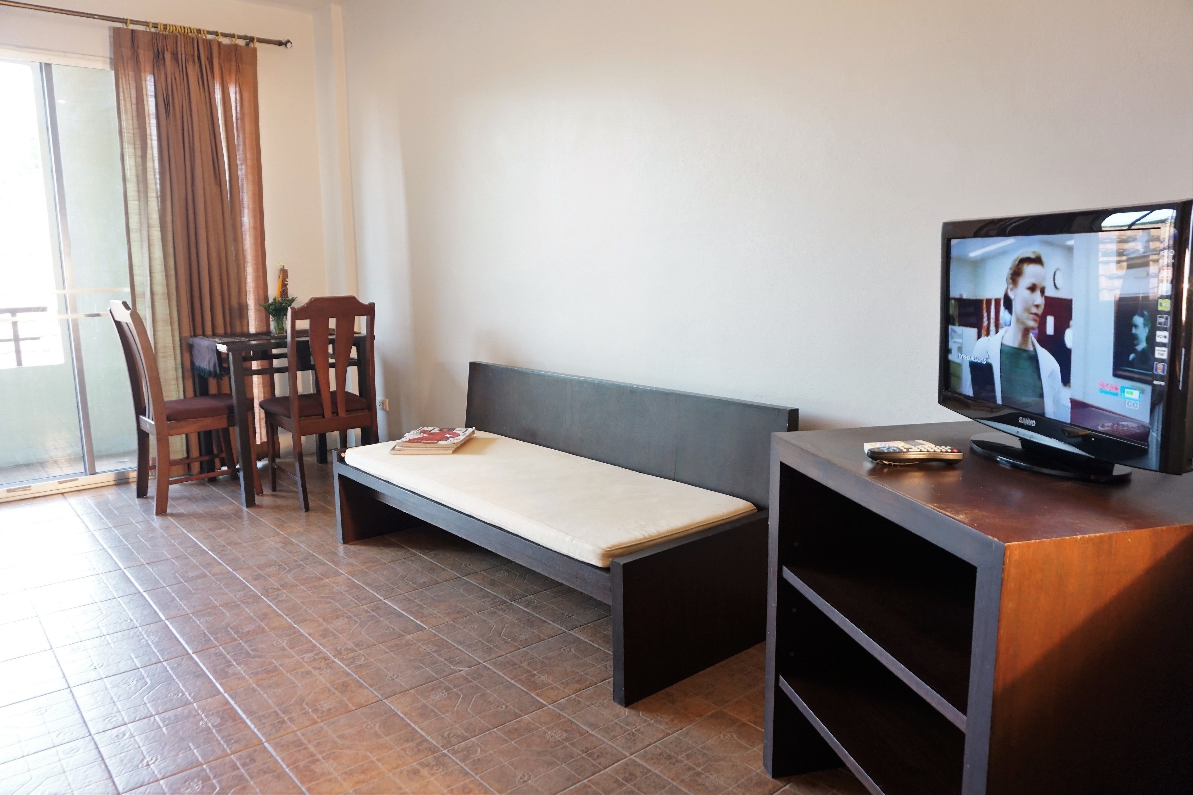 A room with a bed, a TV, a table with chairs, and a window with curtains.