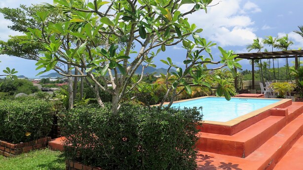 A pool area with steps leading down to the water, surrounded by lush greenery and a clear sky. - Phompassorn Apartment (Chalong)