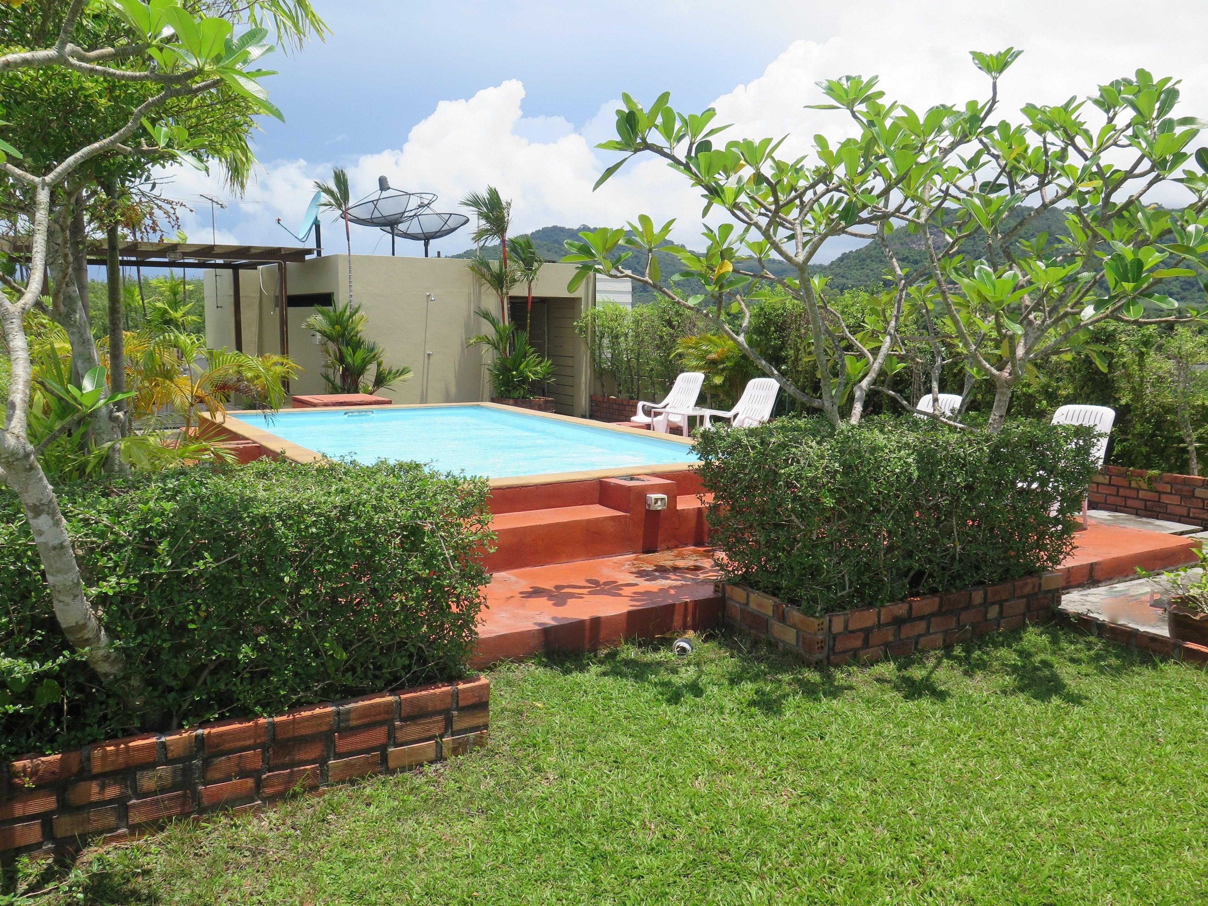 A swimming pool with a brick border, surrounded by grass and plants, and white lounge chairs.