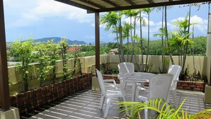 A covered patio with a table and chairs, surrounded by plants and a view of palm trees and distant buildings. - Phompassorn Apartment (Chalong)
