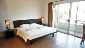 A hotel room with a large bed, a nightstand, a lamp, and a view of the outside through a sliding door. - Phompassorn Apartment (Chalong)