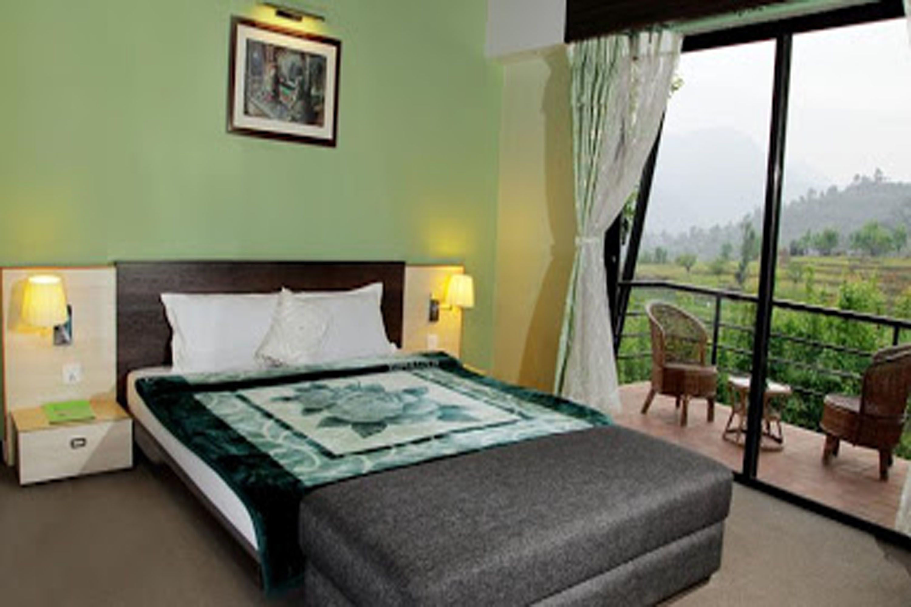 A bedroom with a bed, two bedside lamps, a nightstand, a window with a view of greenery, and a balcony with chairs.