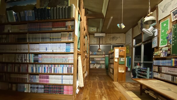 A library with wooden bookshelves filled with books, a wooden floor, and a ceiling fan. - Tomarigi - Hostel (Yakushima)