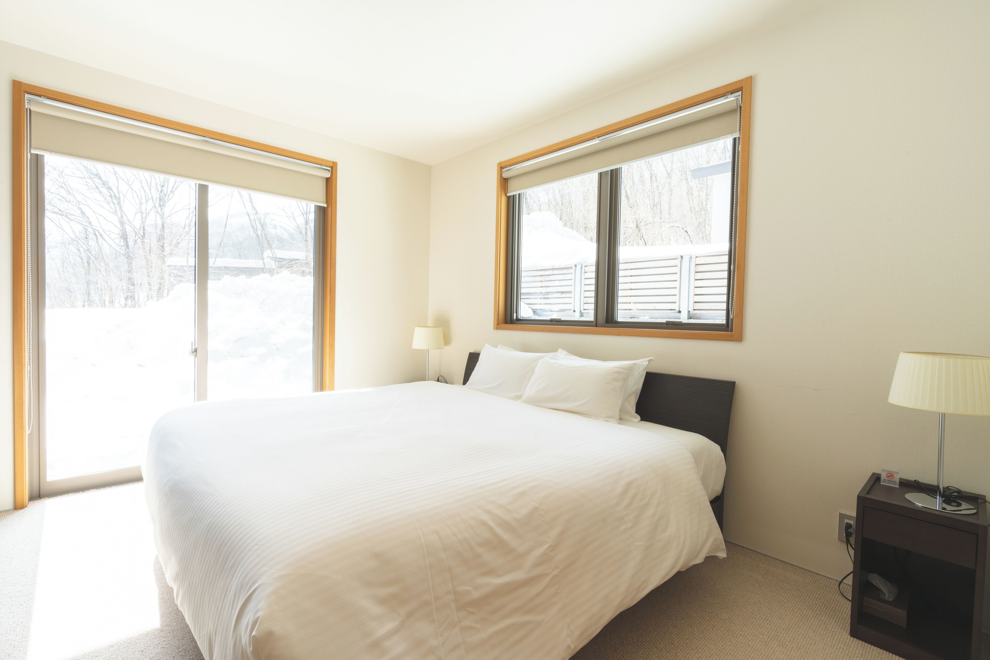 A hotel room with a large bed, two windows with blinds, a bedside table with a lamp, and a view of the outdoors.
