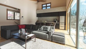 Een moderne woonkamer met een grijze hoekbank, een salontafel en uitzicht op een keuken en buitenruimte door grote ramen.