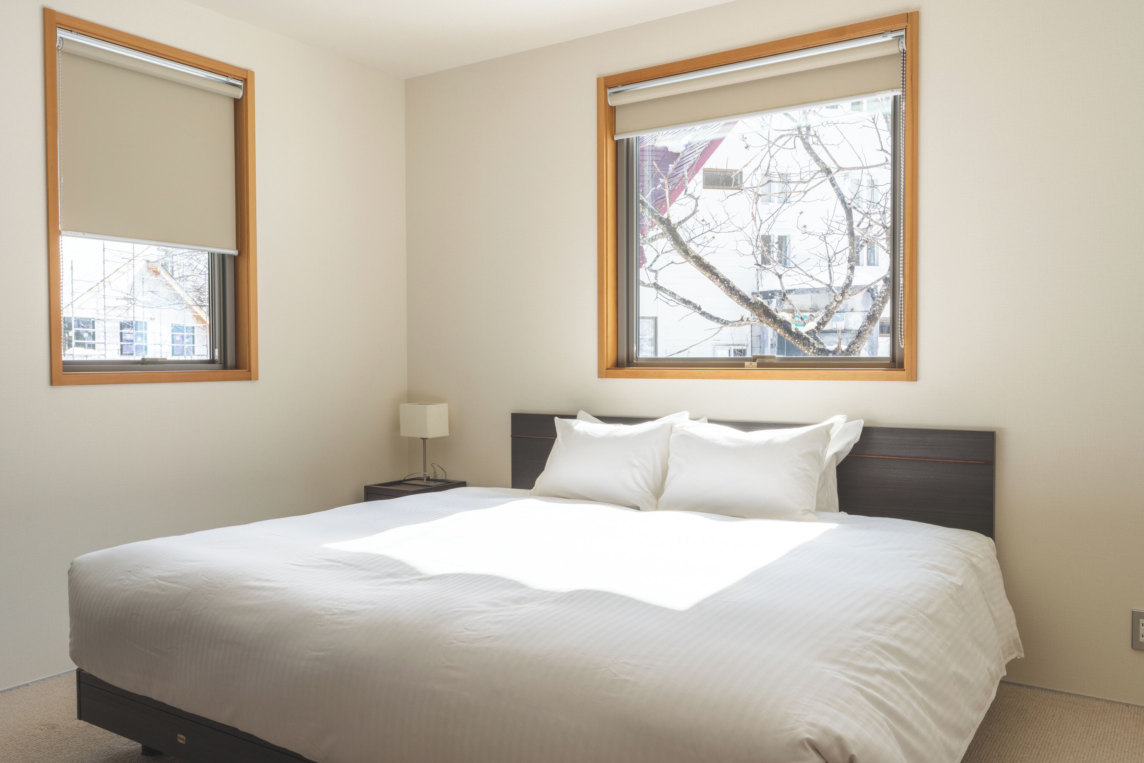 Una cama bien hecha en una habitación de hotel, con un ventanal amplio, una mesita de noche con lámpara y vistas al exterior.