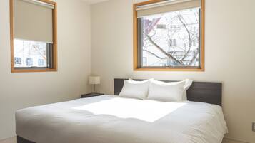 Una cama bien hecha en una habitación de hotel, con un ventanal amplio, una mesita de noche con lámpara y vistas al exterior.