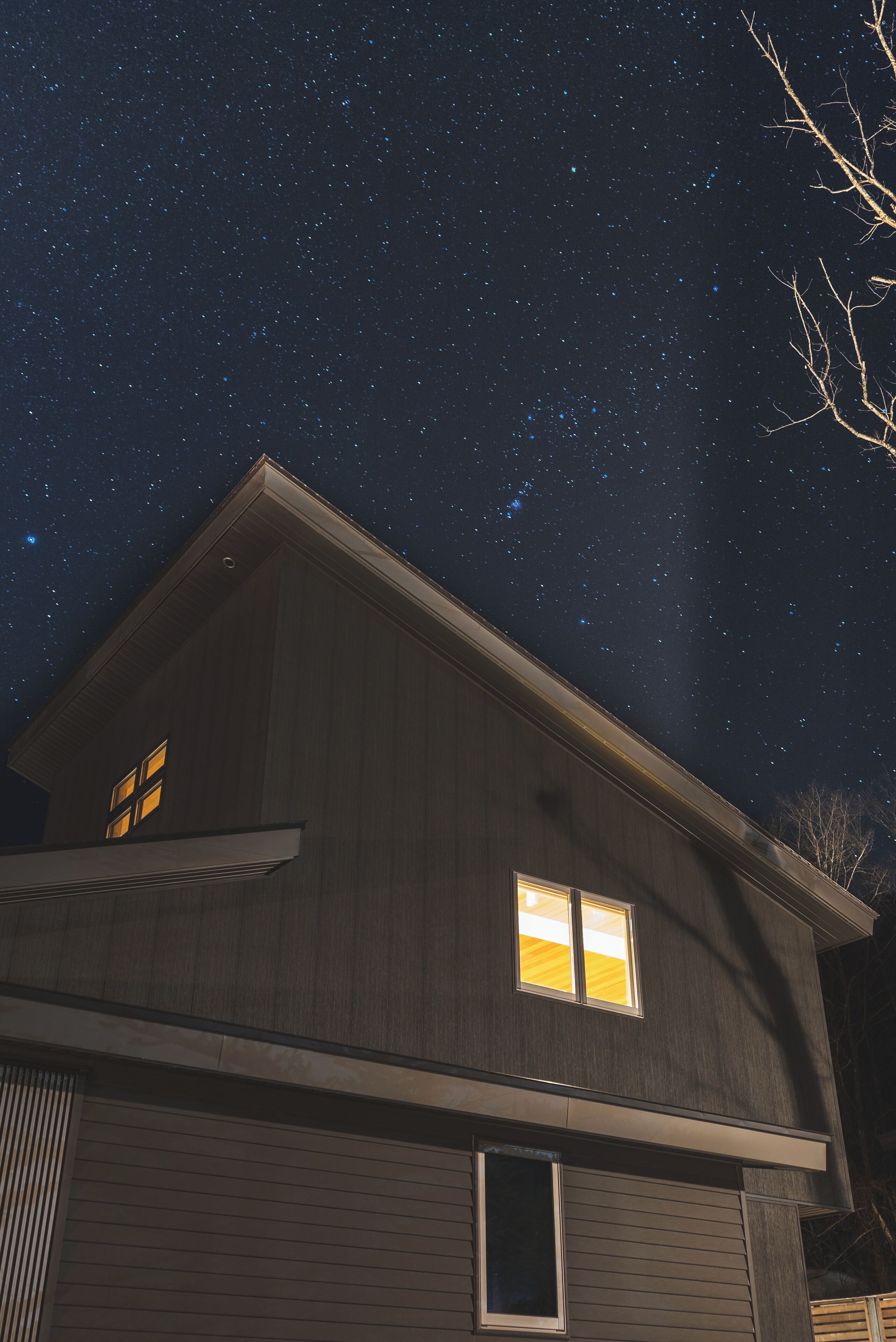 A house with a dark exterior and two lit windows, under a starry night sky.
