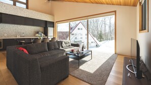 Een moderne woonkamer met een bank, salontafel en televisie. Er is een keukenhoek met een aanrecht en kastjes. Grote ramen bieden uitzicht op het exterieur.