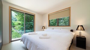 Een groot bed met witte lakens, twee nachtkastjes met lampen en grote ramen met zonwering.